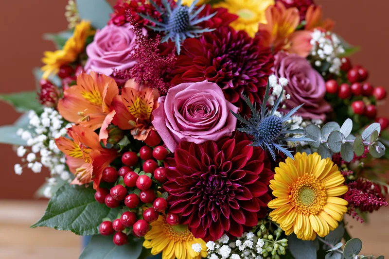 Dense autumn floral bouquet with pink roses, red dahlias, orange alstroemeria, yellow chrysanthemums, thistle, eucalyptus, and red berries arranged in a centerpiece