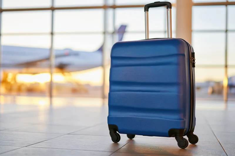 Blue hard-shell rolling suitcase standing upright on tiled floor in airport terminal with airplane visible through window in background