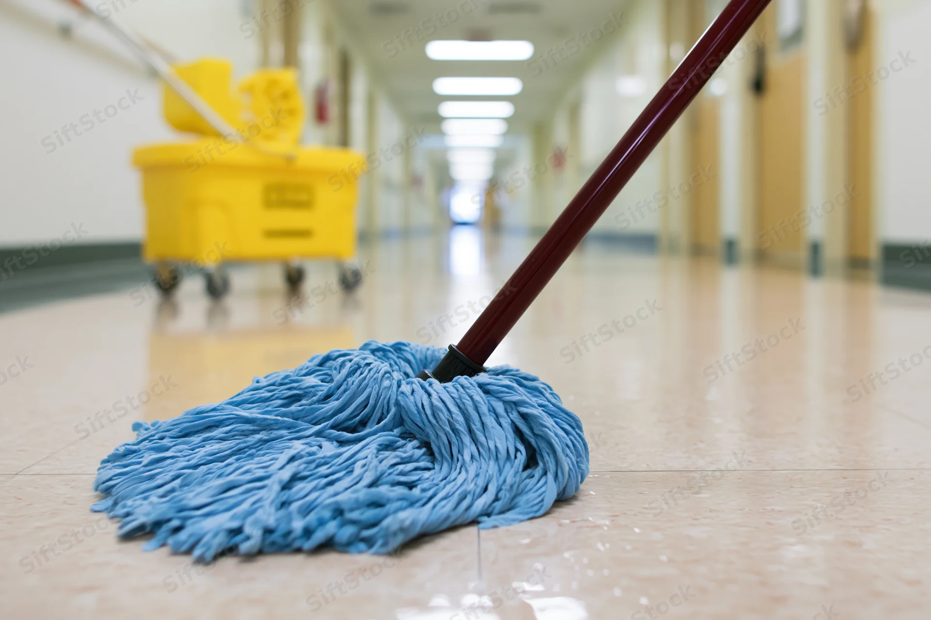 Blue string mop with red handle resting on wet floor in long institutional hallway