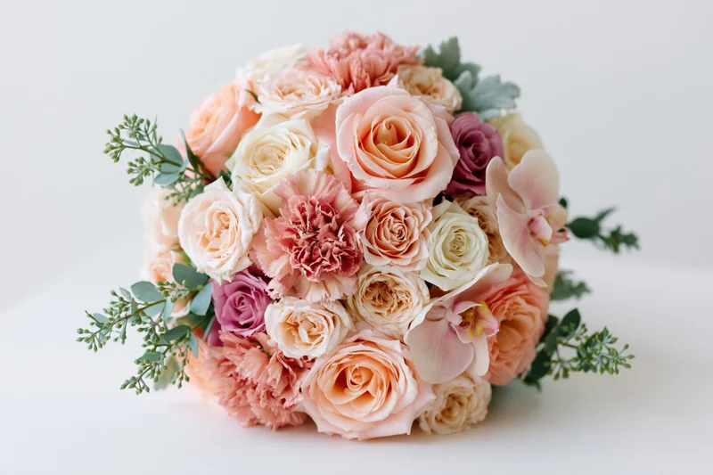 Bridal bouquet densely arranged with roses, orchids, carnations and greenery resting on a flat white surface