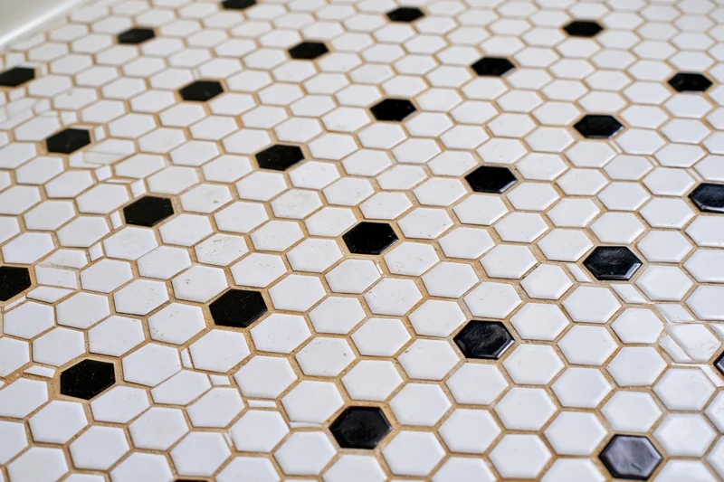 Hexagonal mosaic tile floor with white and black tiles arranged in a repeating pattern, grouted in beige, viewed flat-on