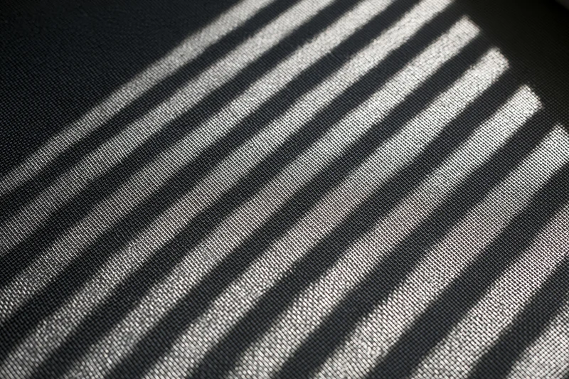 Close-up of dark grey window screen mesh with diagonal light and shadow stripes in flat lay arrangement