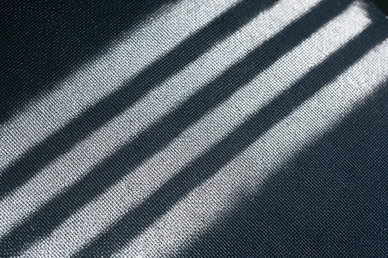 Close-up of dark grey window screen mesh with diagonal light and shadow stripes across its textured surface
