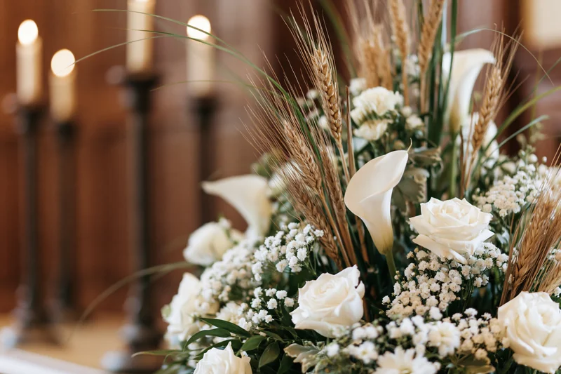 White floral arrangement with roses, calla lilies and wheat on an altar with lit candles in the background