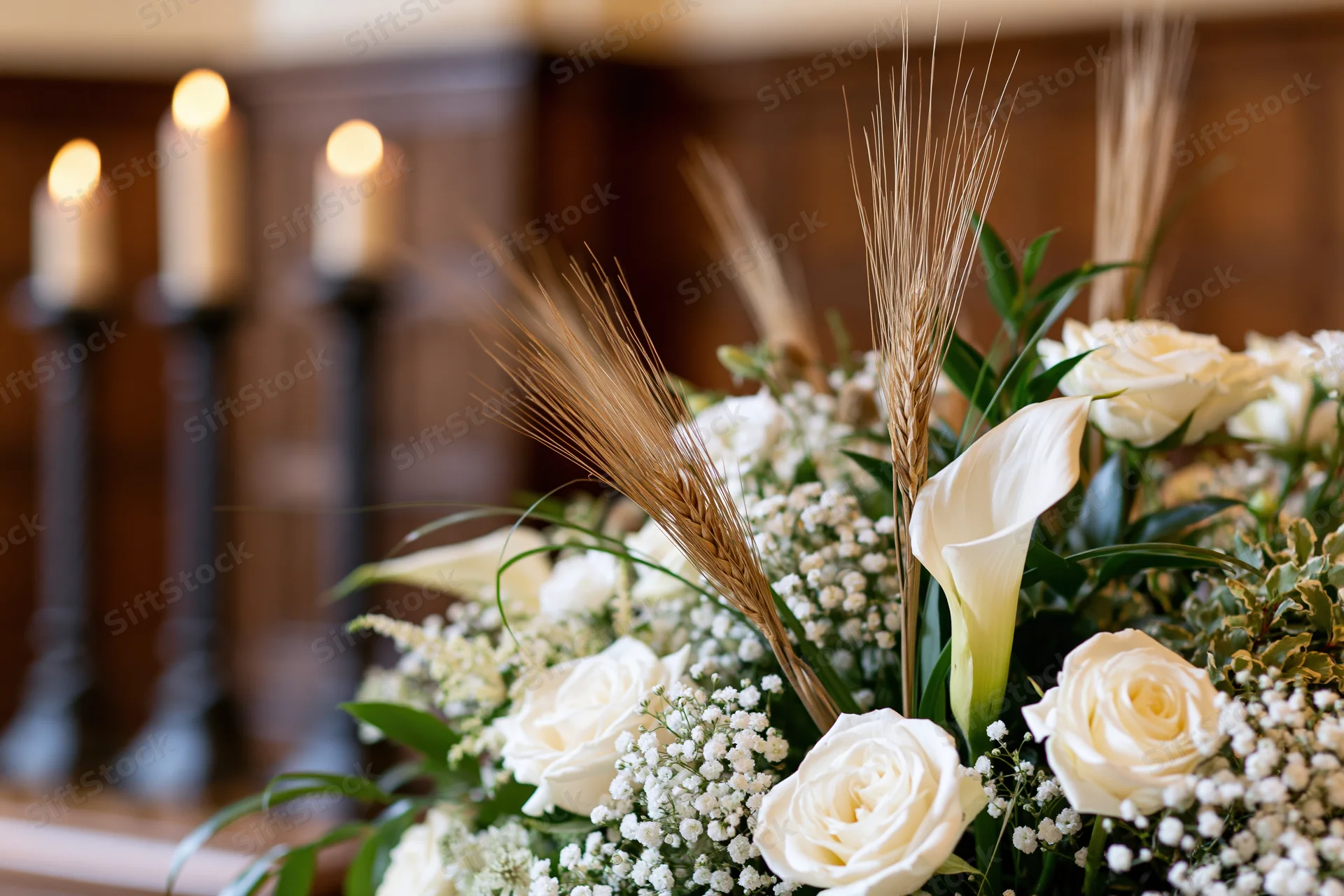 White floral arrangement with roses, calla lilies and wheat stalks on an altar with lit candles in the background