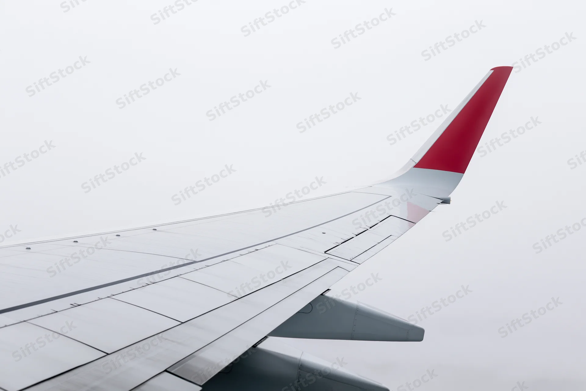 Commercial airplane wing viewed from passenger window with red tail fin against overcast sky