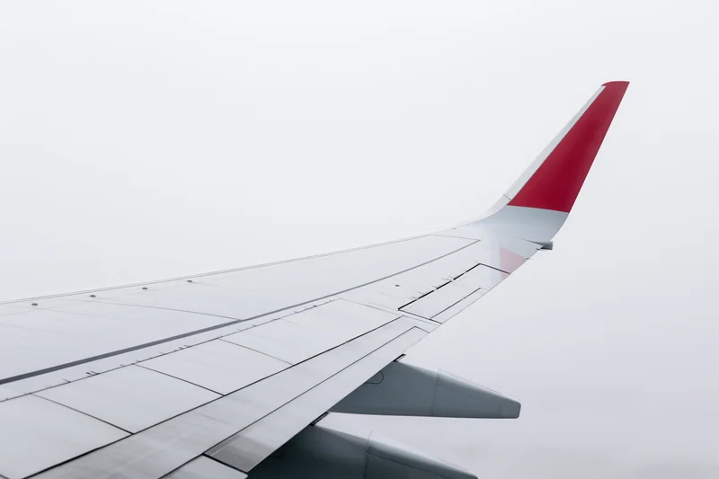 Commercial airplane wing viewed from passenger window with red tail fin against overcast sky