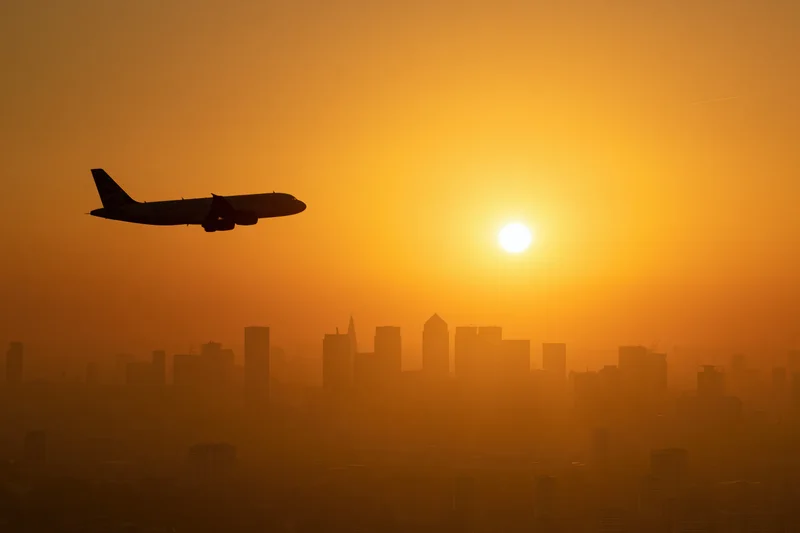 Commercial passenger jet silhouetted against glowing sunset over city skyline