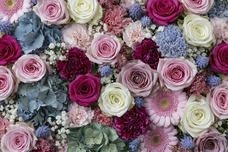 Dense flat-lay arrangement of mixed cut flowers including roses, hydrangeas, carnations, and gerberas in pink, white, purple, and blue hues