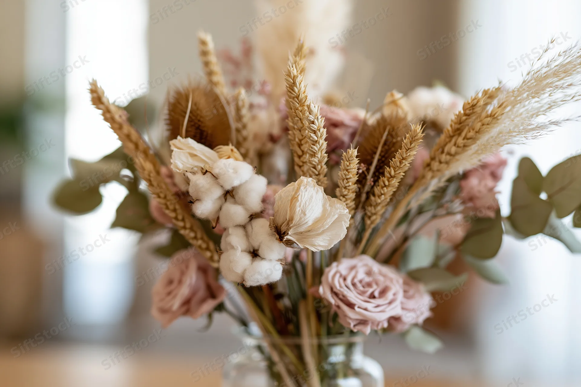 Dried flower arrangement with cotton, wheat, roses and eucalyptus in a glass vase on a table