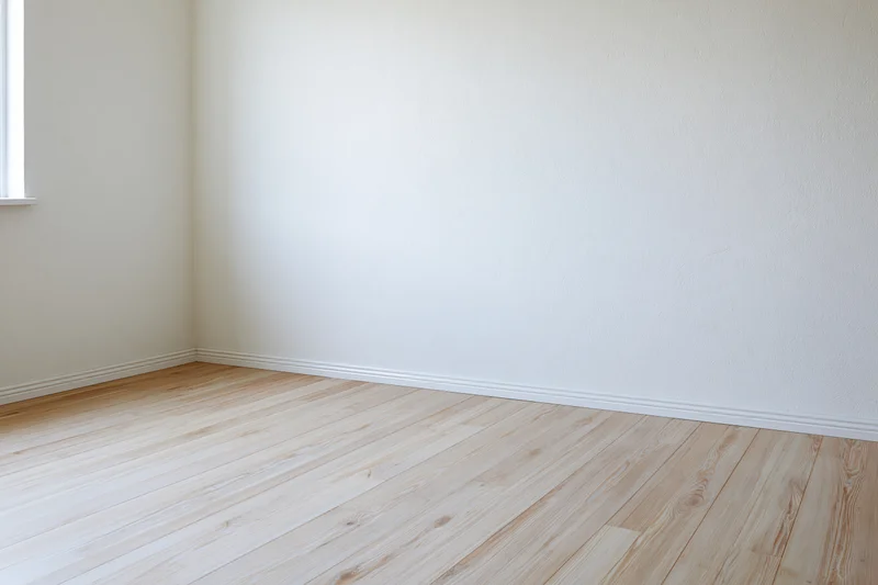 Empty interior corner with off-white plaster walls and light wood flooring, natural light from window
