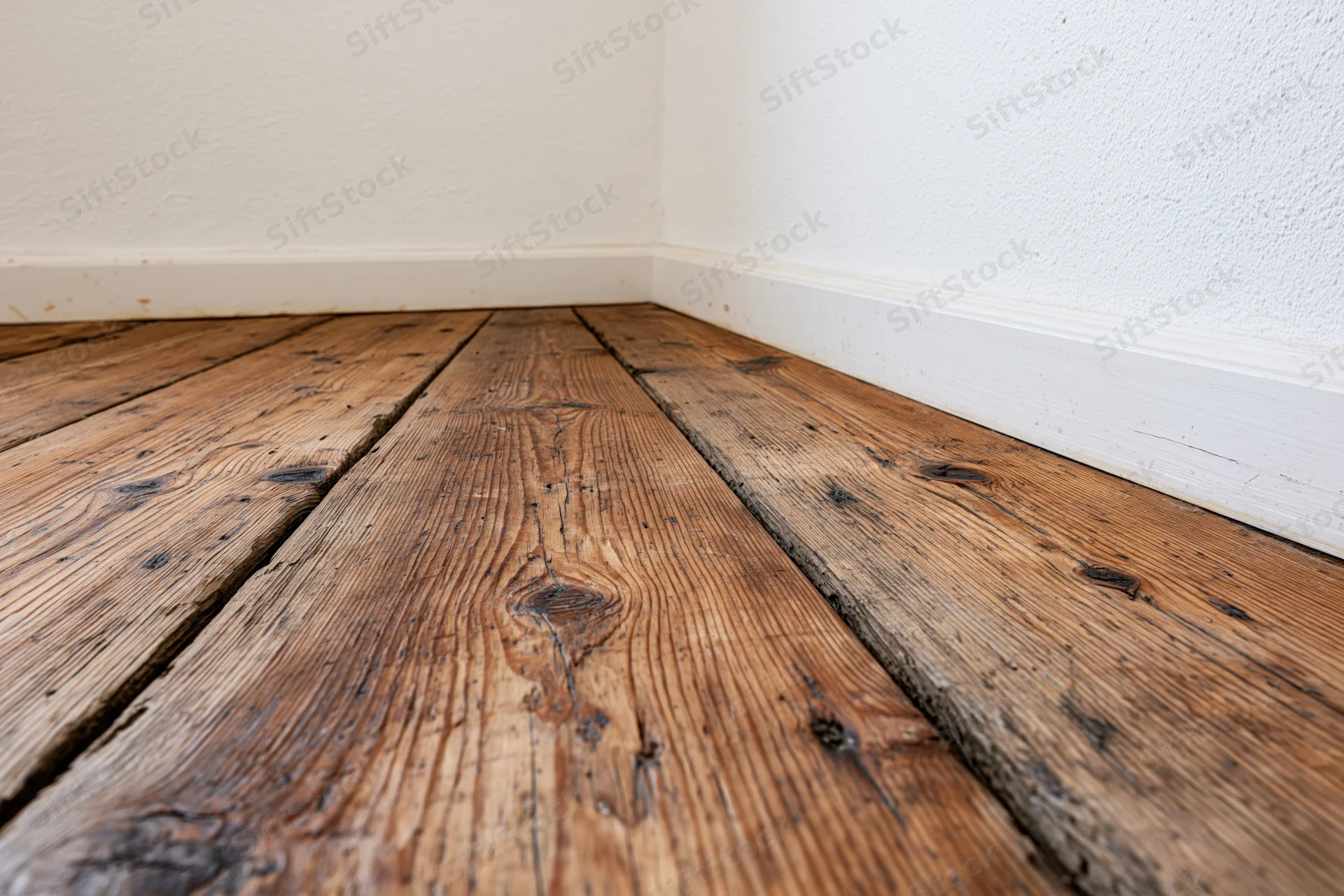 Worn dark brown wooden plank floor in interior corner with white walls and baseboard