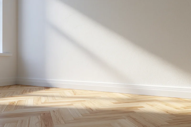 Empty interior room with white walls and light herringbone wood flooring in corner near window