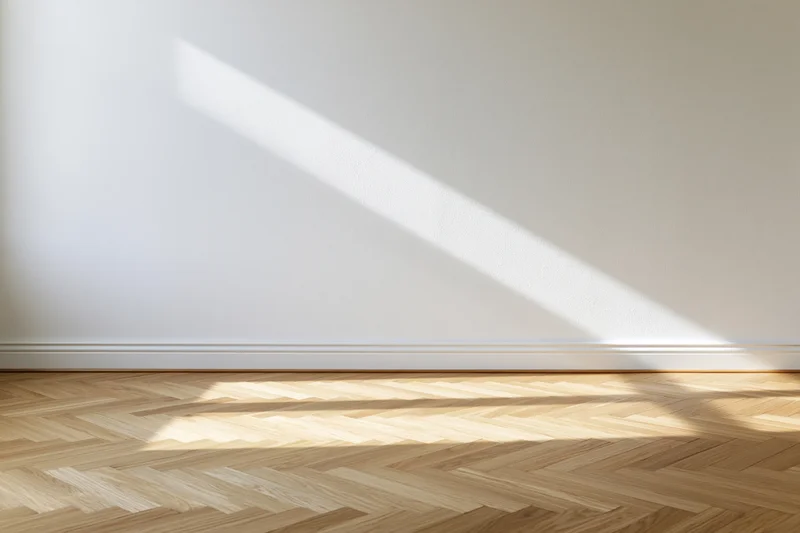 Empty room with white wall and light herringbone wood floor, sunlight casting diagonal shadow across surface
