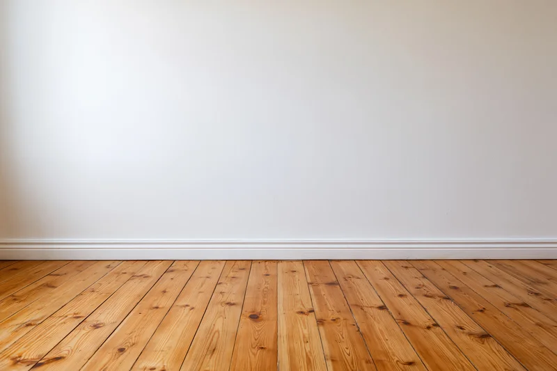 Empty room with plain white wall and bare wooden floor showing baseboard and wood grain texture