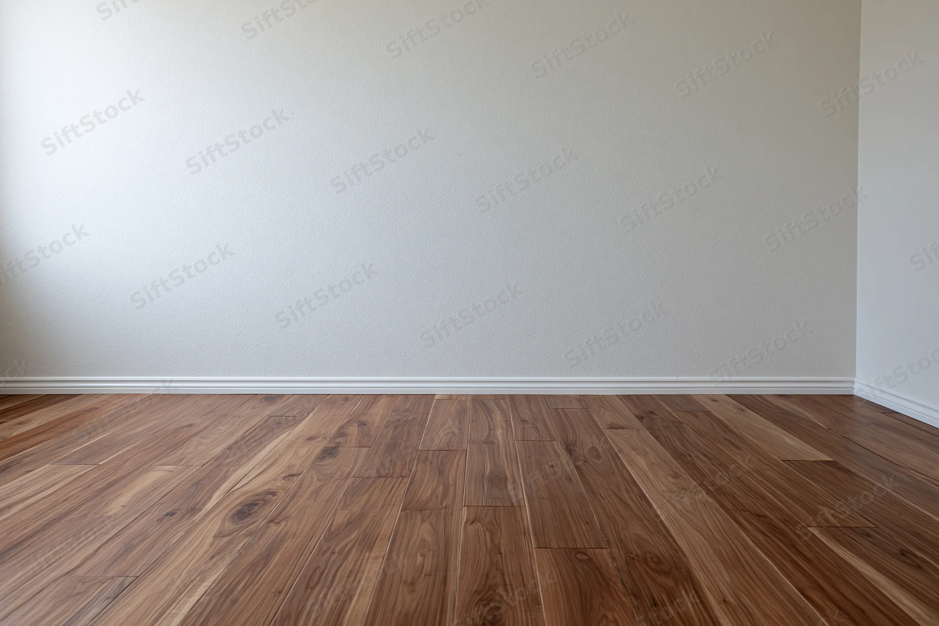 Empty interior room with white wall and medium-toned wood flooring meeting at baseboard