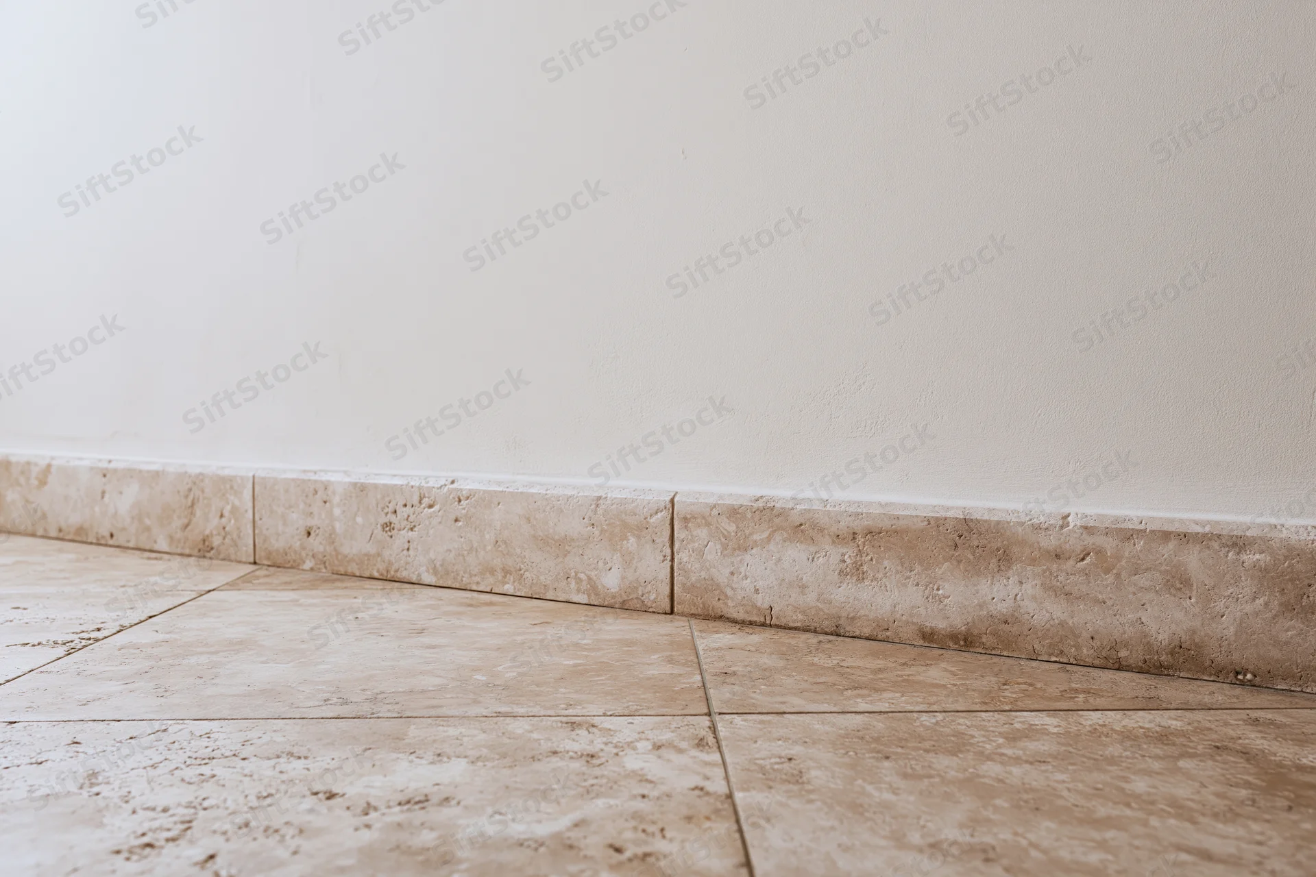 Beige tiled floor meeting white plaster wall with baseboard trim in empty interior space