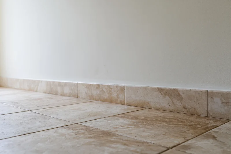 Beige tiled floor meeting white plaster wall with baseboard trim in empty room