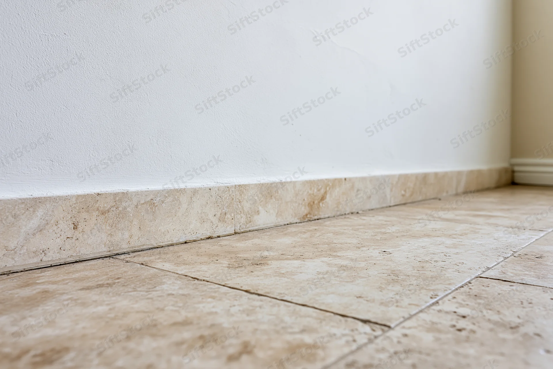 Beige travertine tile flooring meeting white plaster wall at corner with baseboard trim