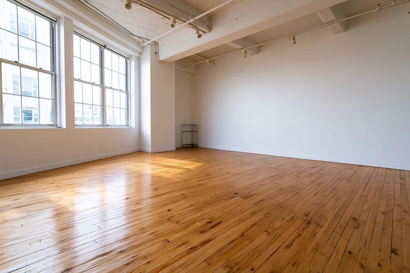 Empty loft studio space with white walls and hardwood floor, featuring large windows and exposed ceiling beams