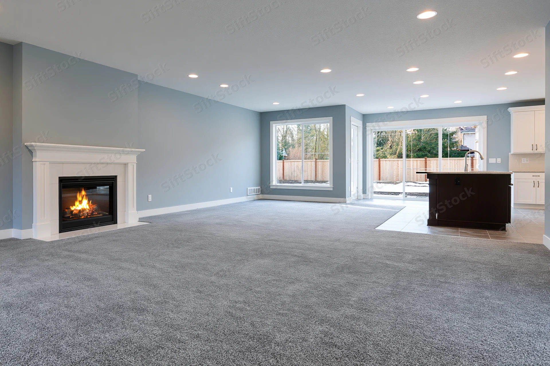 Empty open-plan living room with lit fireplace and kitchen island visible through sliding door to patio