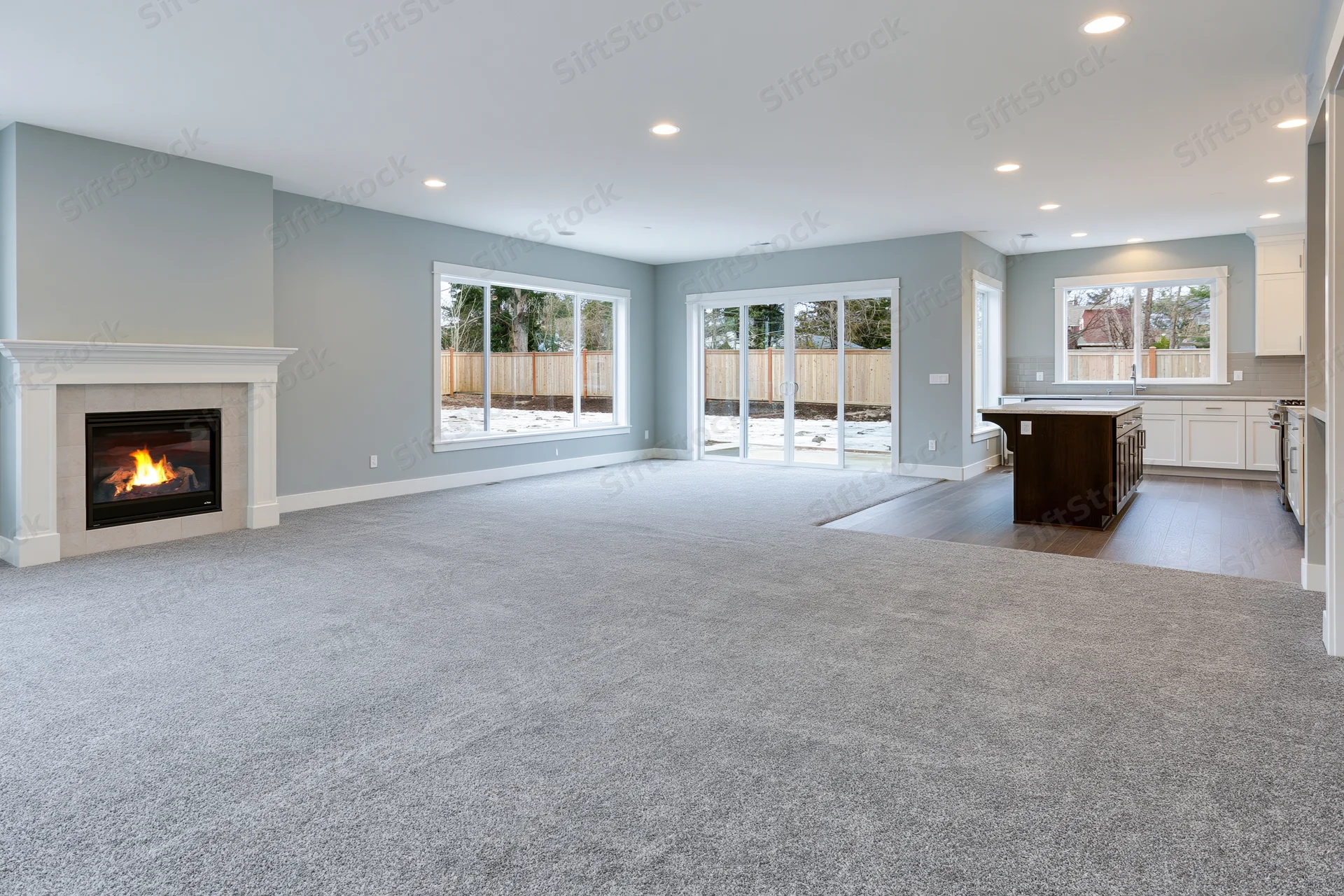 Empty open-plan living room with lit fireplace and kitchen island, featuring gray carpet and large windows overlooking snowy yard
