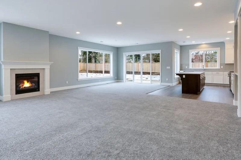 Empty open-plan living room with lit fireplace and kitchen island, featuring gray carpet and large windows overlooking snowy yard