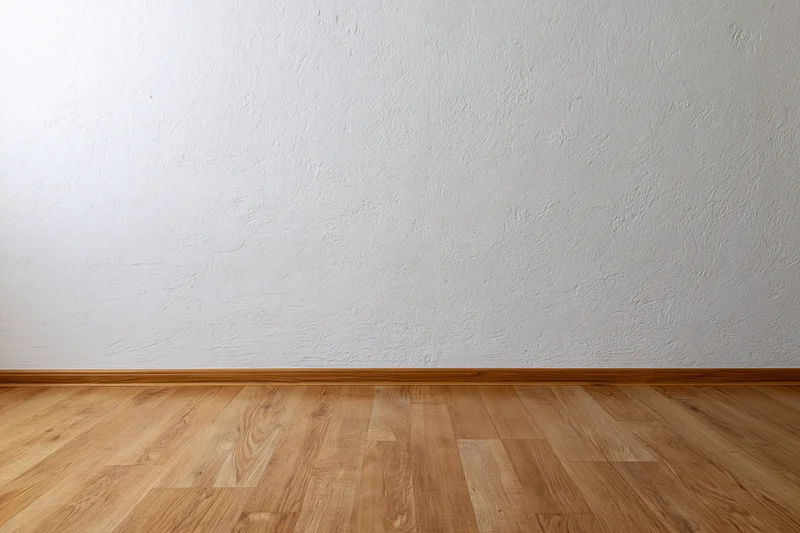 Empty room interior with textured white plaster wall meeting light wood laminate floor at baseboard trim