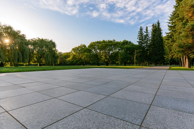 Wide granite tile plaza in a green park with trees and open lawn under morning sunlight