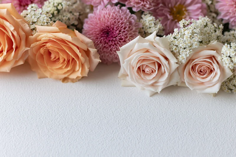 Flat lay of peach roses, pink chrysanthemums and white sprays arranged on textured white surface with copyspace