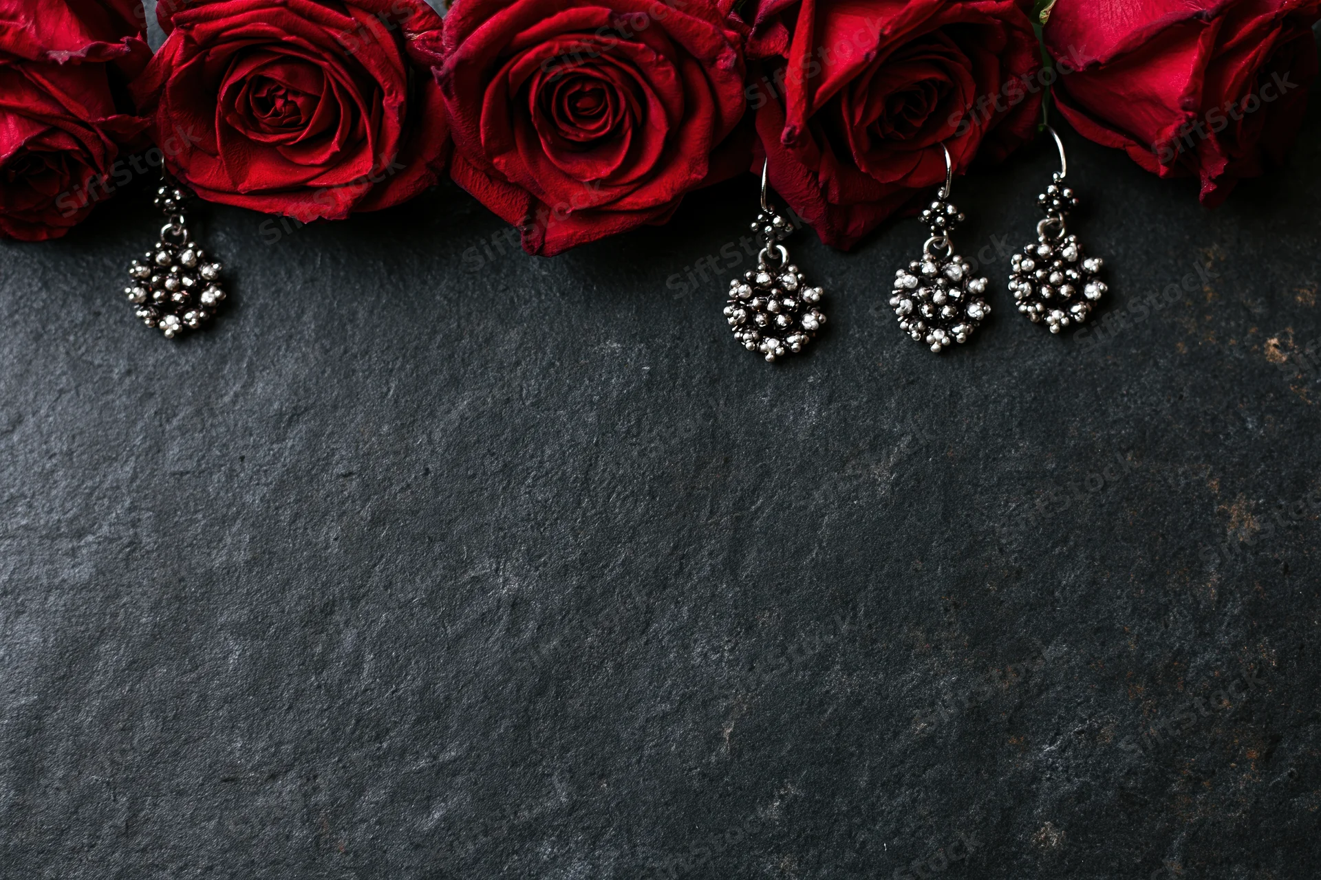 Deep crimson roses and ornate silver cluster earrings arranged in a flat lay on a dark textured stone surface with copyspace below