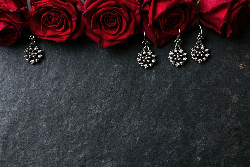 Deep crimson roses and ornate silver cluster earrings arranged in a flat lay on a dark textured stone surface with copyspace below