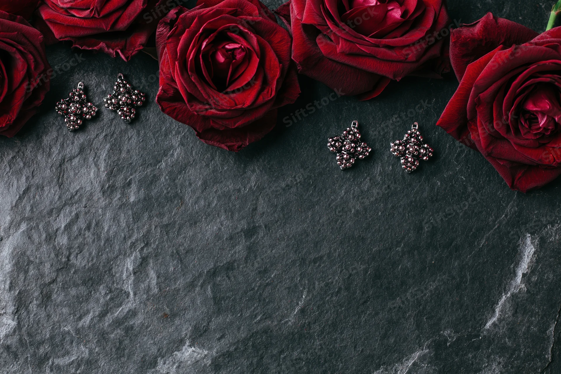 Deep crimson roses and ornate silver cluster earrings arranged in flat lay on dark stone surface with copyspace