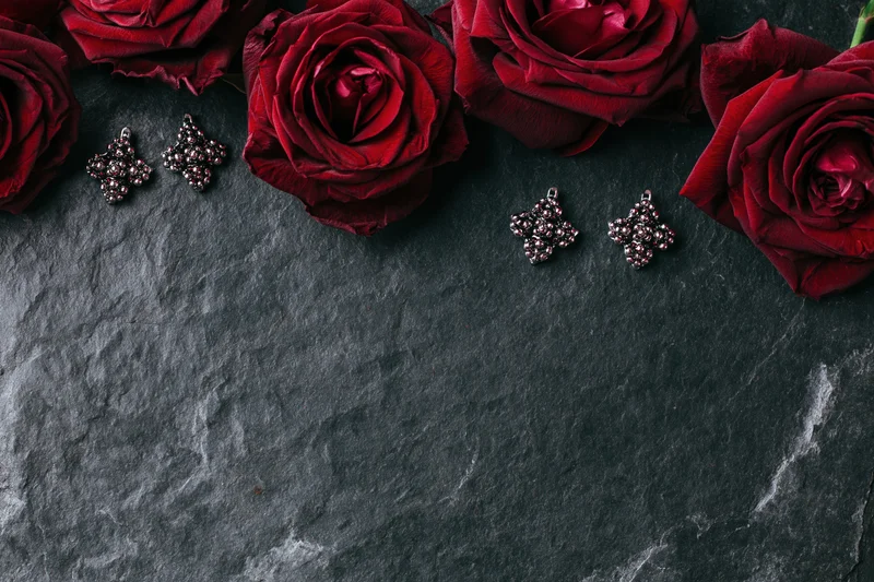 Deep crimson roses and ornate silver cluster earrings arranged in flat lay on dark stone surface with copyspace