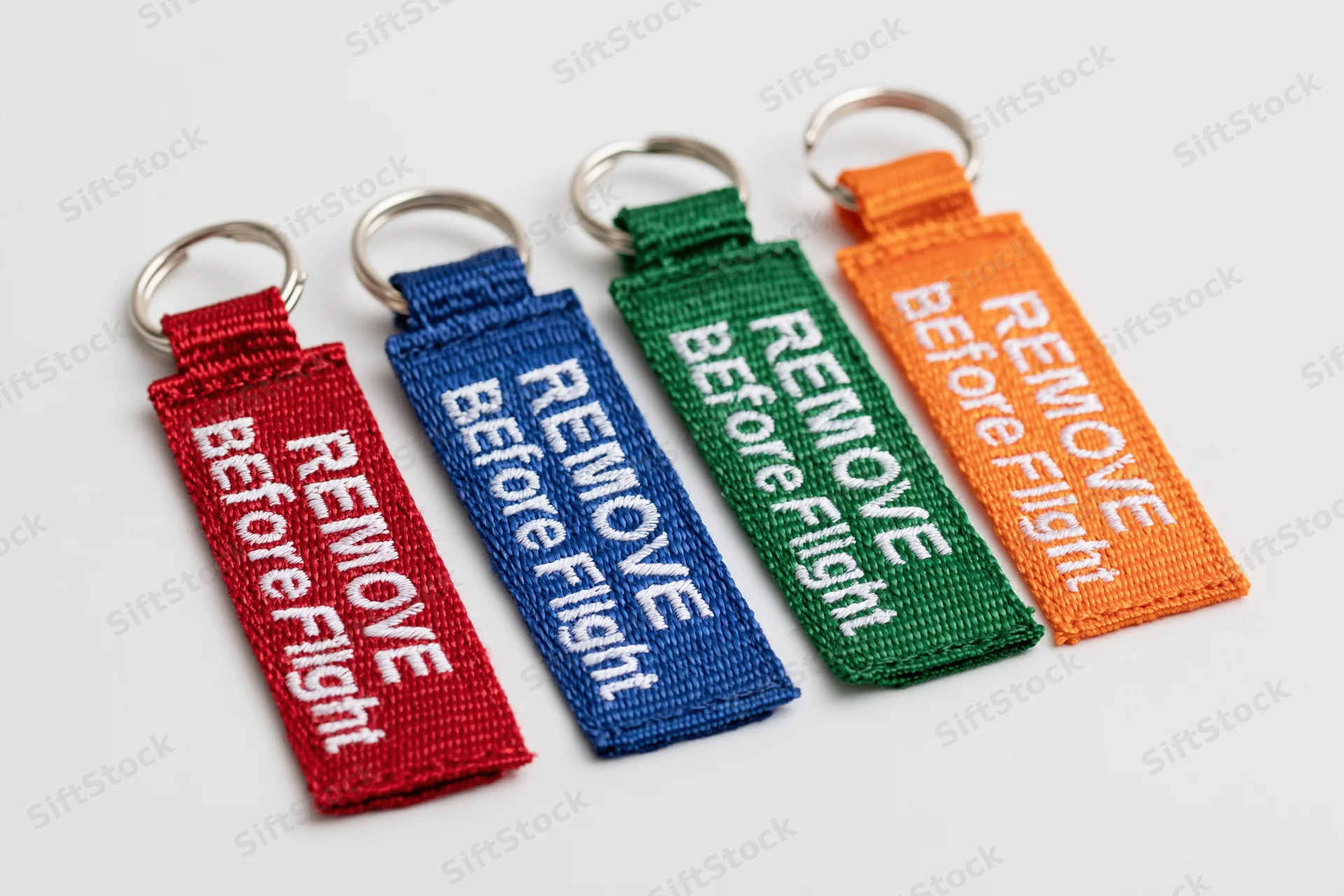 Four embroidered fabric keychain tags reading remove before flight in red blue green and orange arranged in a row on a white surface