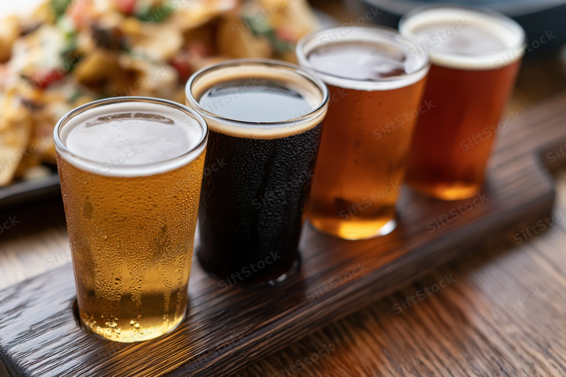 Four craft beer tasting glasses arranged on a dark wooden paddle with condensation and froth, placed on a wooden table