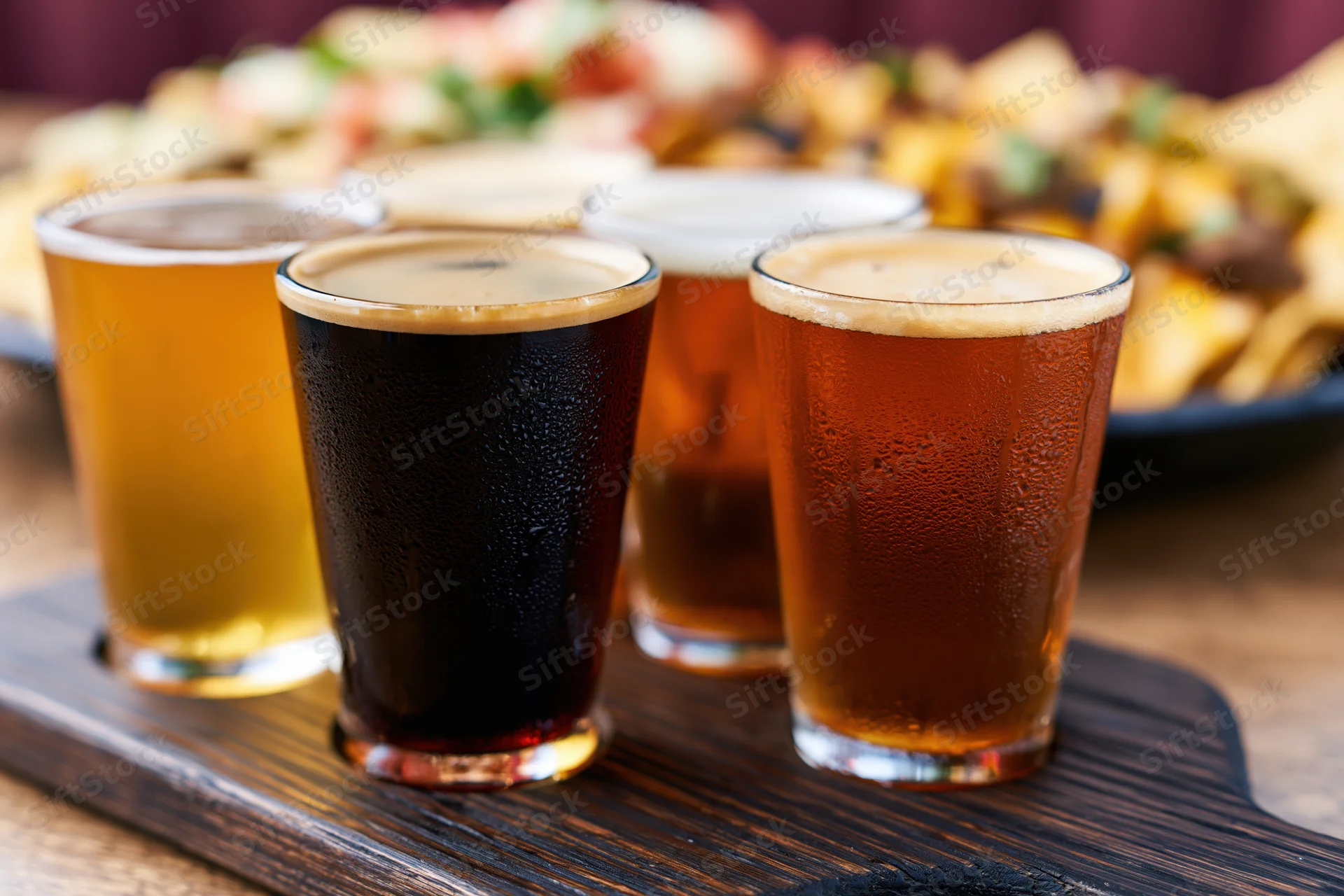 Four craft beer tasting flight glasses arranged on a dark wooden paddle with condensation and foam, served with snacks in the background