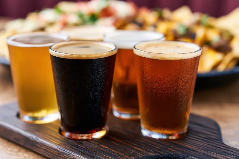 Four craft beer tasting flight glasses arranged on a dark wooden paddle with condensation and foam, served with snacks in the background