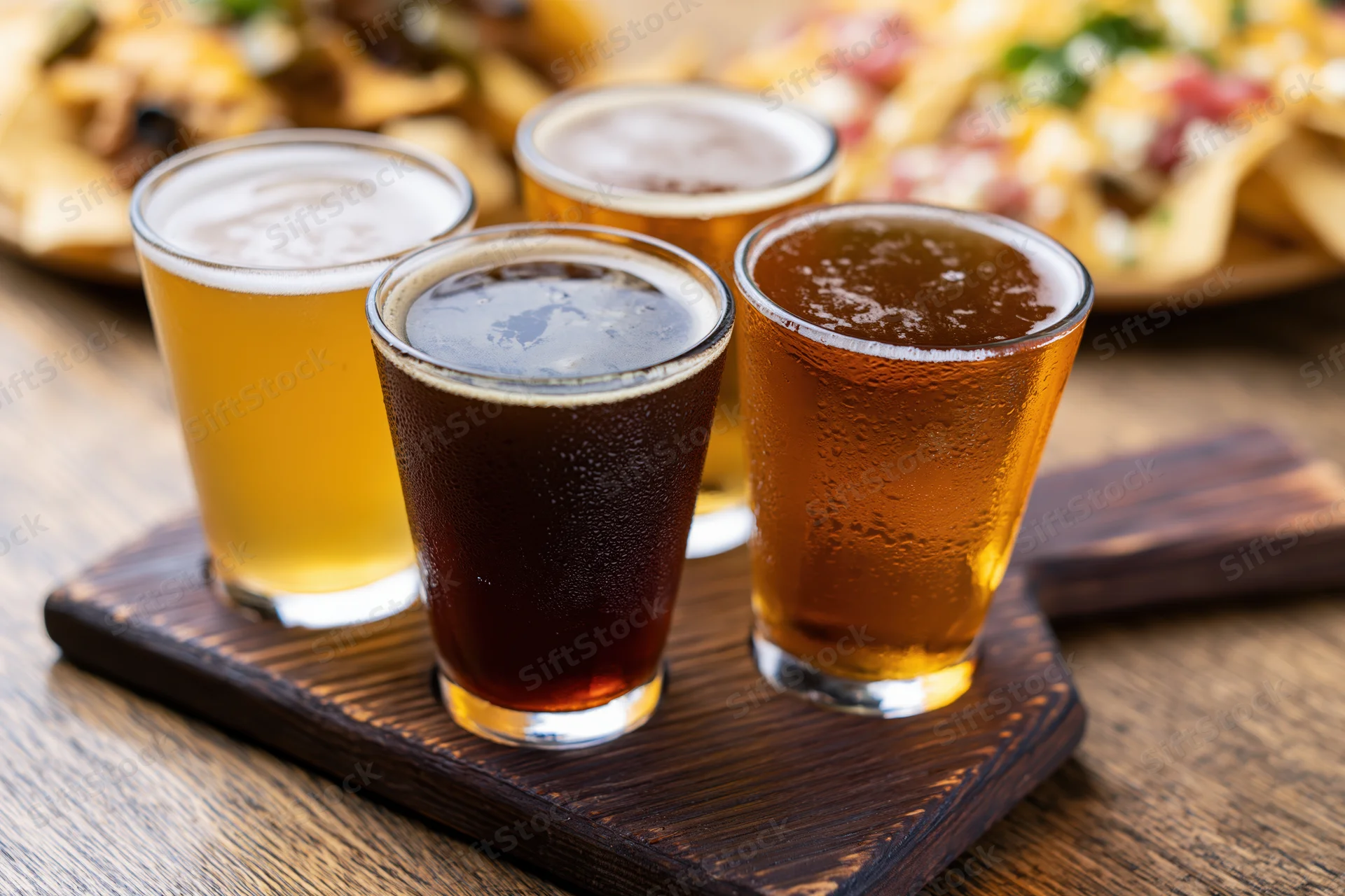 Four craft beer tasting flight glasses arranged on a dark wooden paddle with condensation and froth, accompanied by nachos in the background