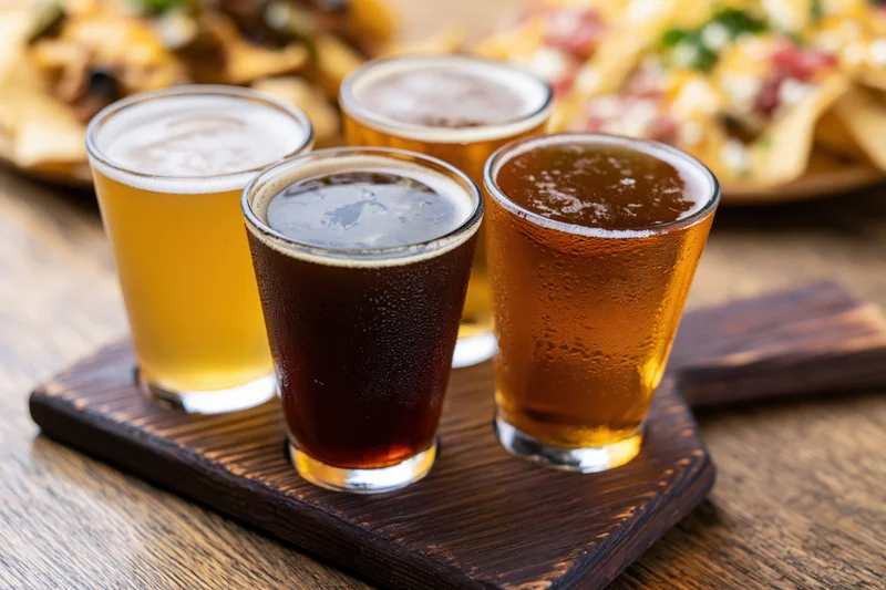 Four craft beer tasting flight glasses arranged on a dark wooden paddle with condensation and froth, accompanied by nachos in the background