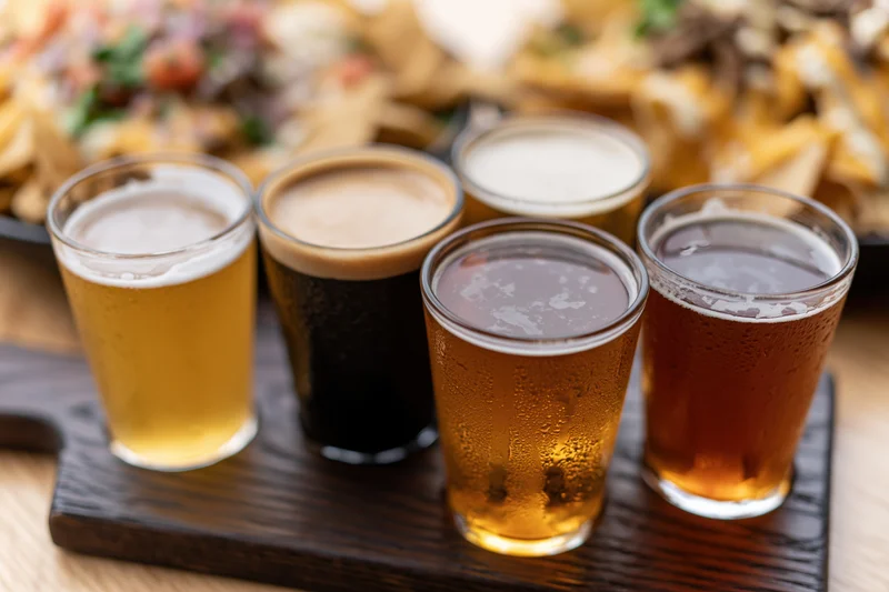 Four craft beer tasting flight glasses arranged on a dark wooden paddle with snacks in background