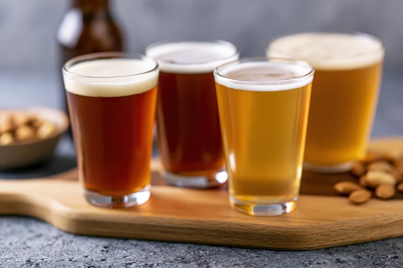 Four craft beer tasting glasses arranged in a row on a wooden tray with peanuts and a bottle in the background