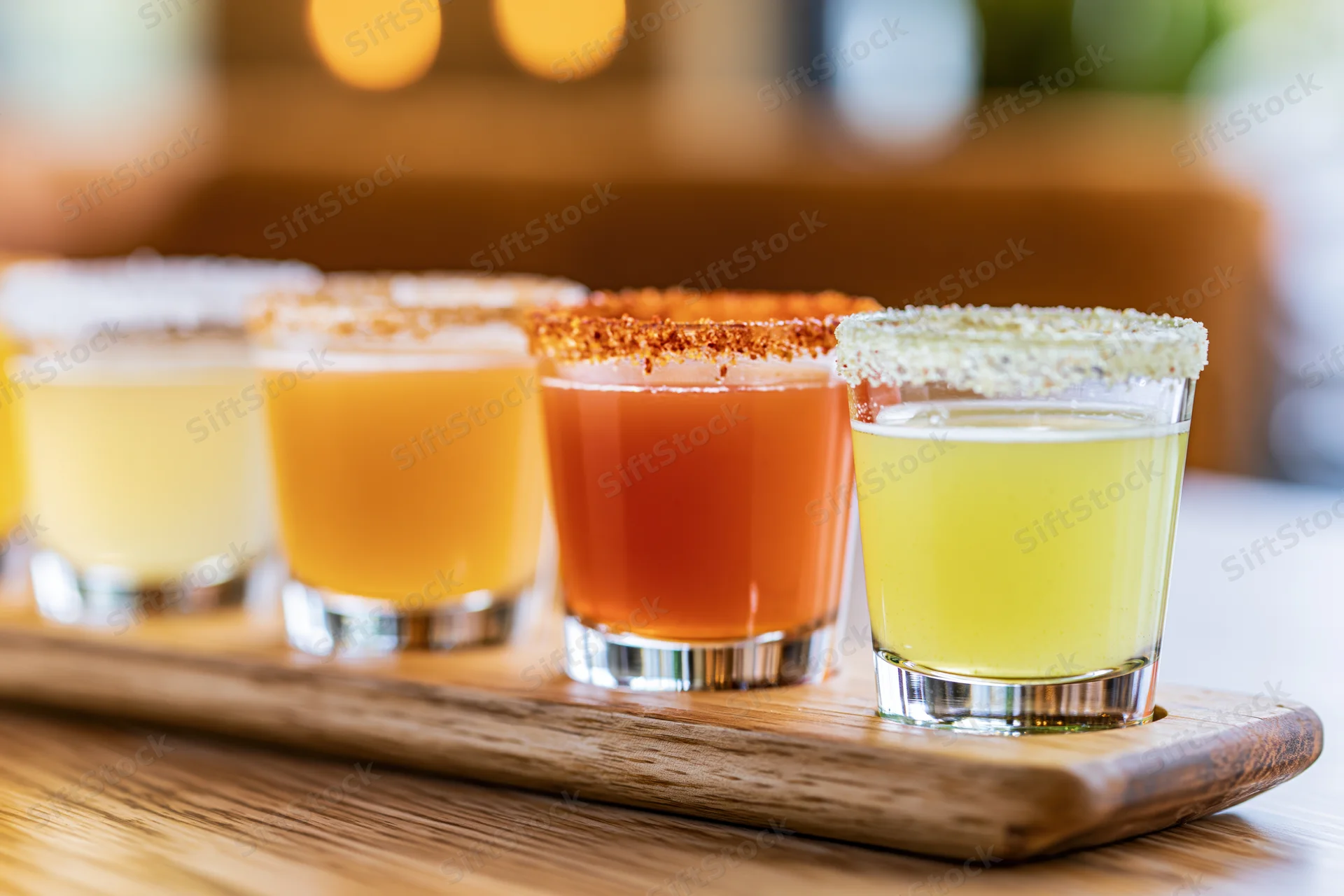 Four margarita flight glasses arranged in a row on a wooden tray with salted and spiced rims, each filled with different colored cocktails