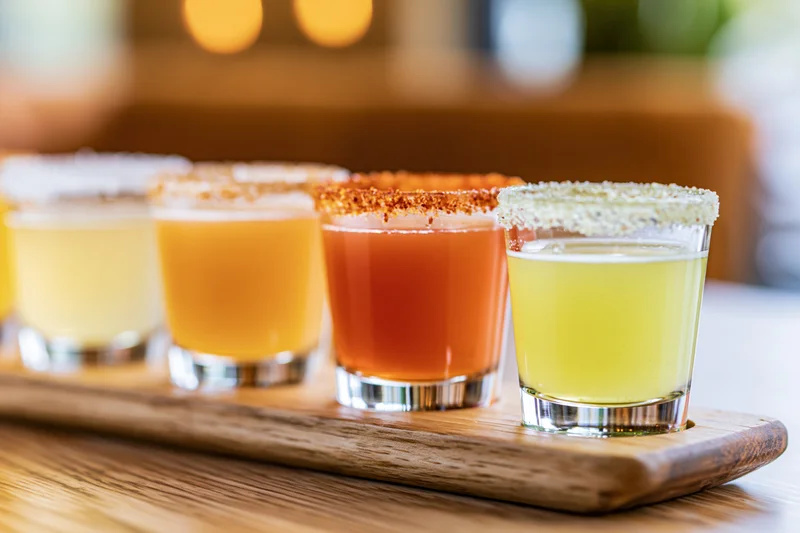 Four margarita flight glasses arranged in a row on a wooden tray with salted and spiced rims, each filled with different colored cocktails