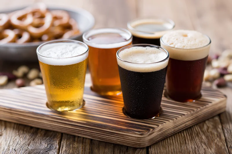 Four small tasting glasses of beer arranged in a row on a wooden tray with snacks nearby