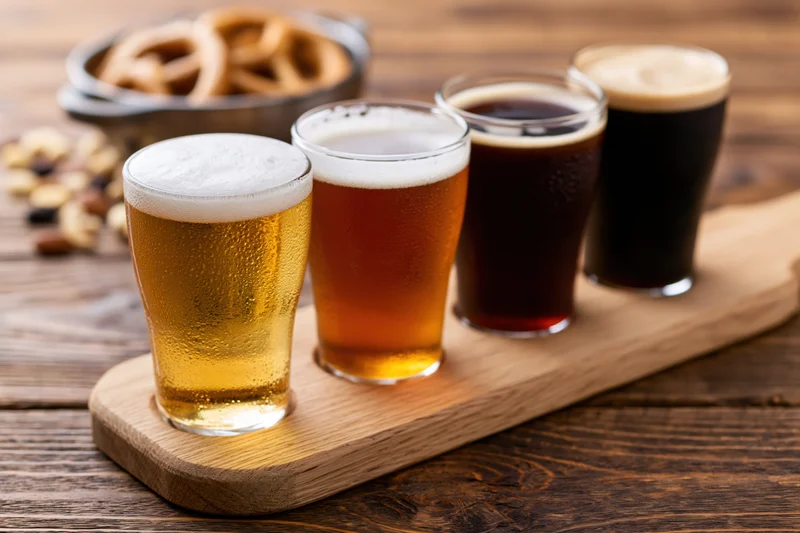 Four small tasting glasses of beer arranged in a row on a wooden tray with pretzels and nuts nearby
