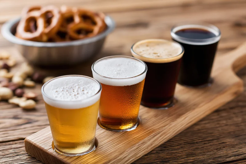 Four small glasses of beer arranged in a row on a wooden tray with pretzels and nuts nearby