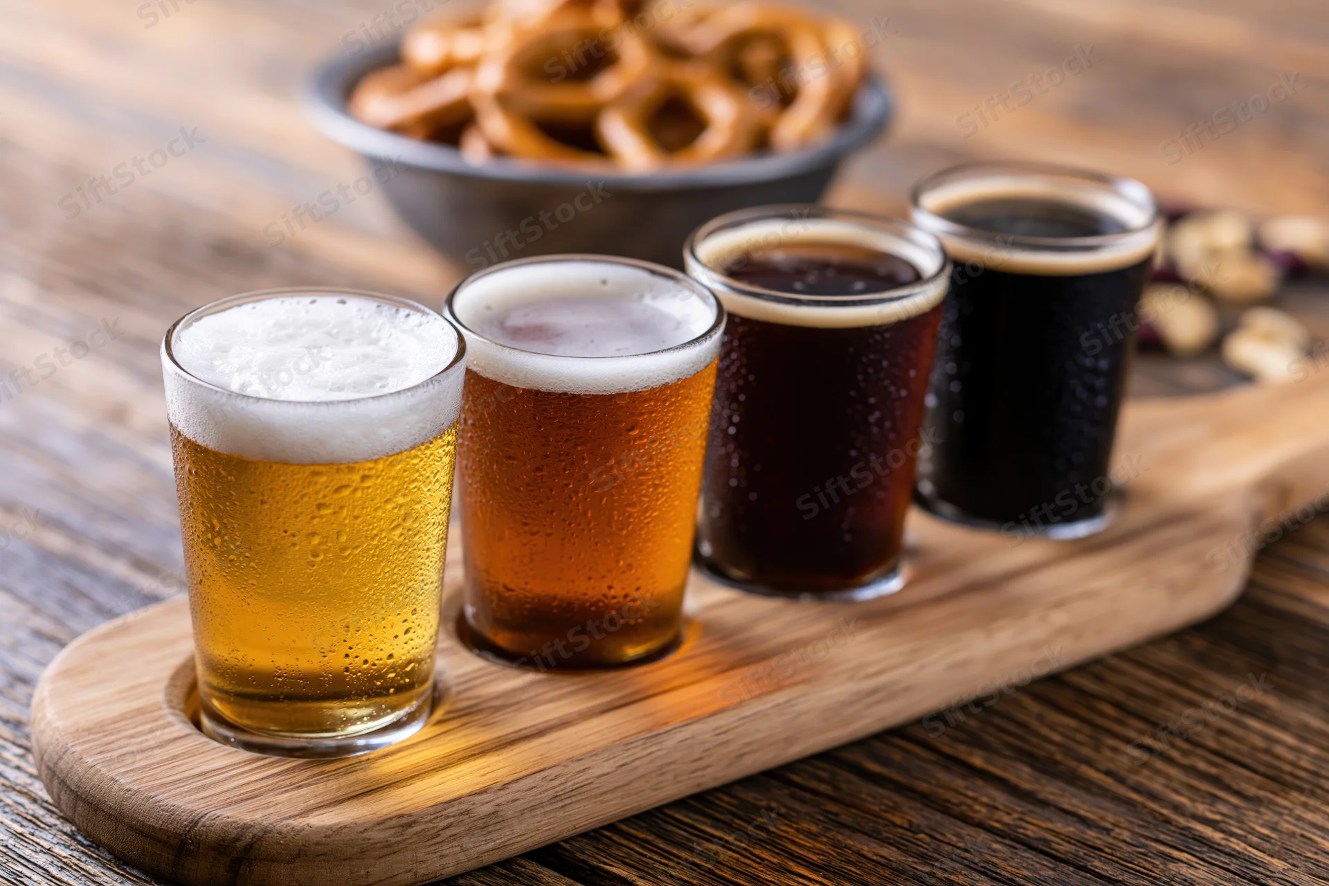 Four small glasses of beer arranged in a row on a wooden tray with condensation and foam, accompanied by pretzels and nuts on a rustic table