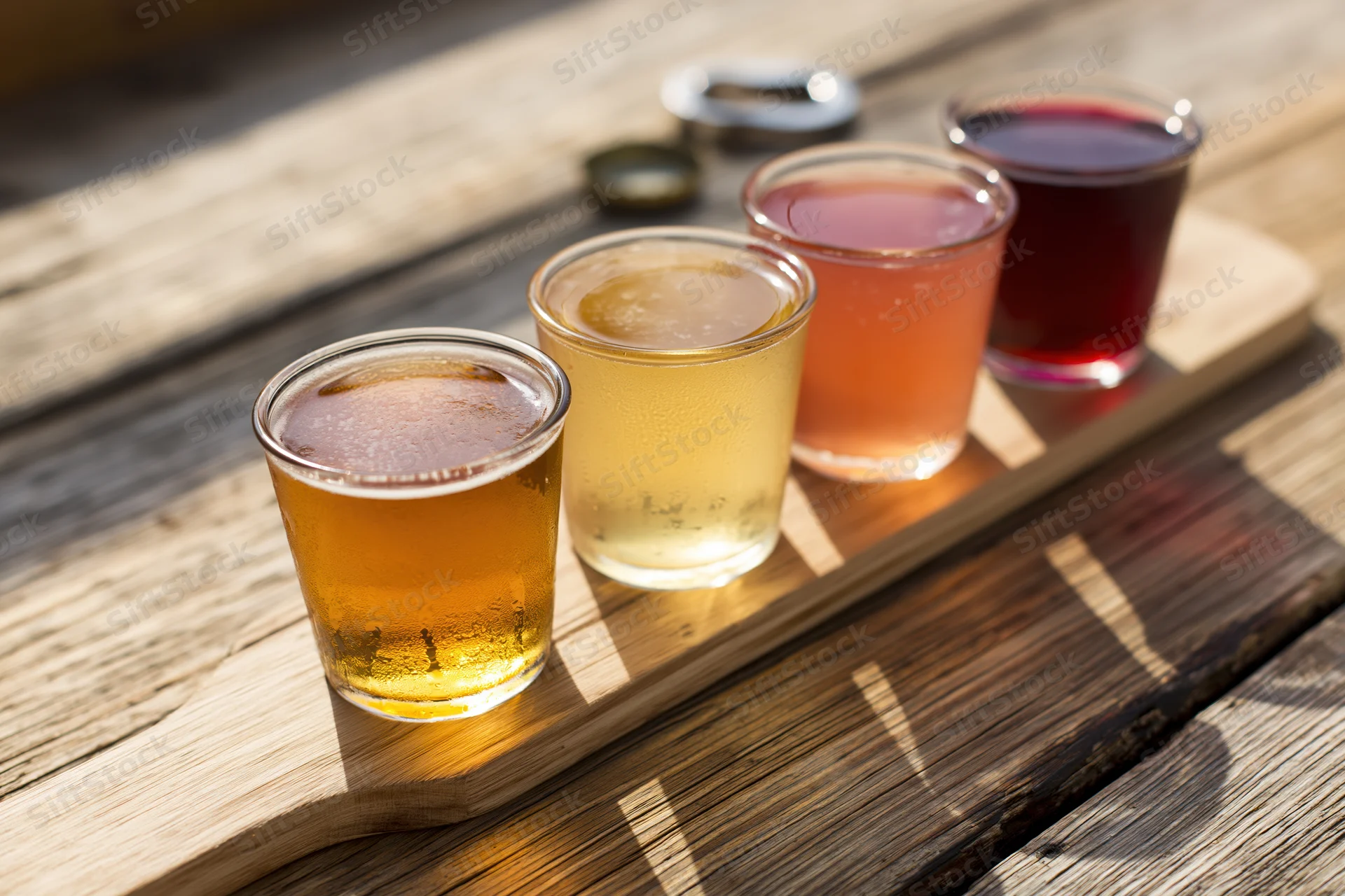 Four small glasses of cider arranged in a row on a wooden tray outdoors in sunlight