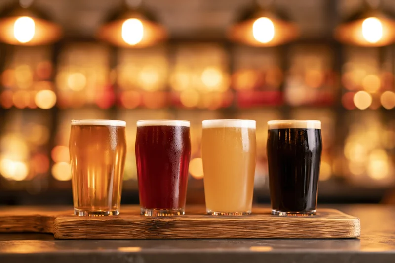 Four craft beer tasting glasses arranged in a row on a wooden tray in a bar setting with warm bokeh lighting in the background